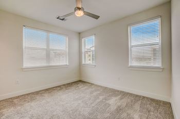 A room with a carpeted floor, a ceiling fan, and two windows with blinds.